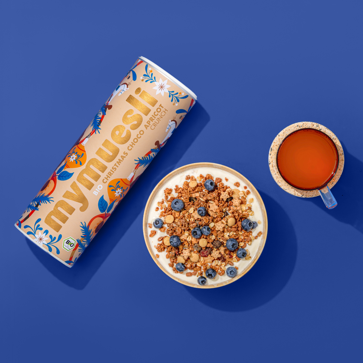 Produktabbildung des mymuesli Christmas Choco Almond Cinnamon Crunch im weihnachtlichen Dosen Design. Dazu eine mit Joghurt angerichtete Müslischale mit frischen Blaubeeren und ein Tee. Alles auf blauem Hintergund.