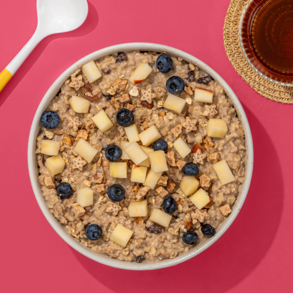 Draufsicht einer Müsli-Schale mit vielen Apfelstücken und Blaubeeren auf einem leuchtend pinken Hintergrund.