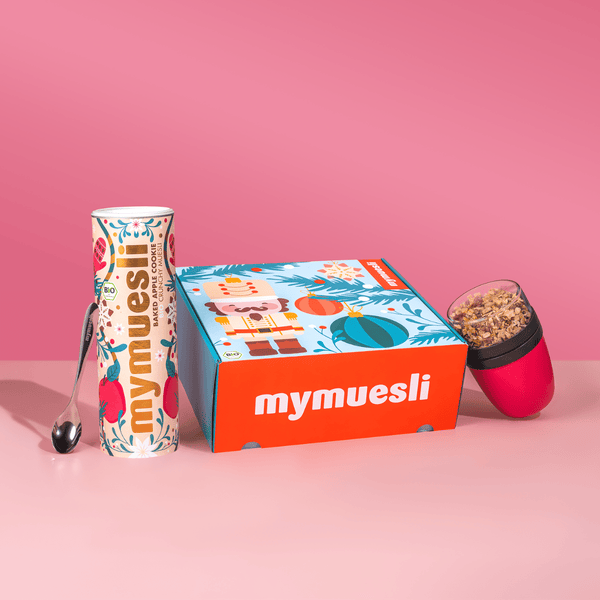 Das mymuesli Geschenkset, bestehend aus Bio Baked Apple Cookie Crunchy, Müsli To-go Becher in magenta / pink und einem silbernen mymuesli Löffel. Verpackt in einer weihnachtlichen Geschenkbox. 