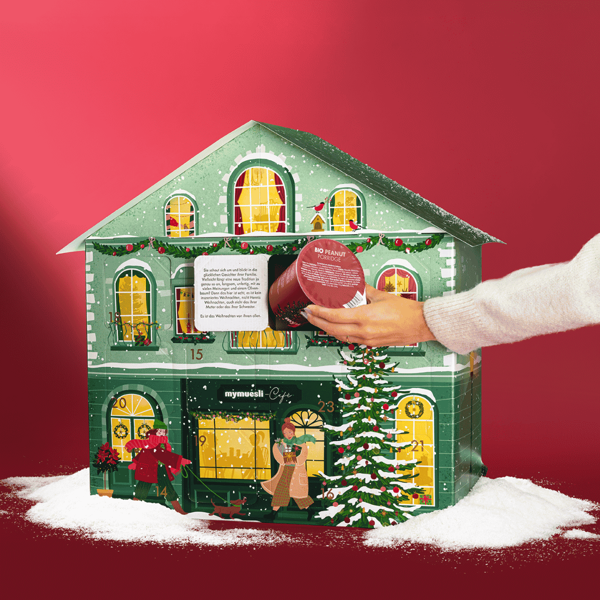 Grünes Adventskalenderhaus mit leichtem Schnee. Vor dem Haus läuft eine Frau mit ihrem Dackel vorbei. Ein Mann steht auf der Leiter und schmückt einen Tannenbaum. Die Fenster des Hauses sind beleuchtet. Eine Frau nimmt einen Müslibecher aus einem geöffneten. 