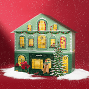 Grünes Adventskalenderhaus mit leichtem Schnee und weihnachtlicher Dekoration. Vor dem Haus läuft eine Frau mit ihrem Dackel vorbei. Ein Mann steht auf der Leiter und schmückt einen Tannenbaum. Die Fenster des Hauses sind beleuchtet. 