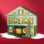 Grünes Adventskalenderhaus mit leichtem Schnee. Vor dem Haus läuft eine Frau mit ihrem Dackel vorbei. Ein Mann steht auf der Leiter und schmückt einen Tannenbaum. Die Fenster des Hauses sind beleuchtet. 