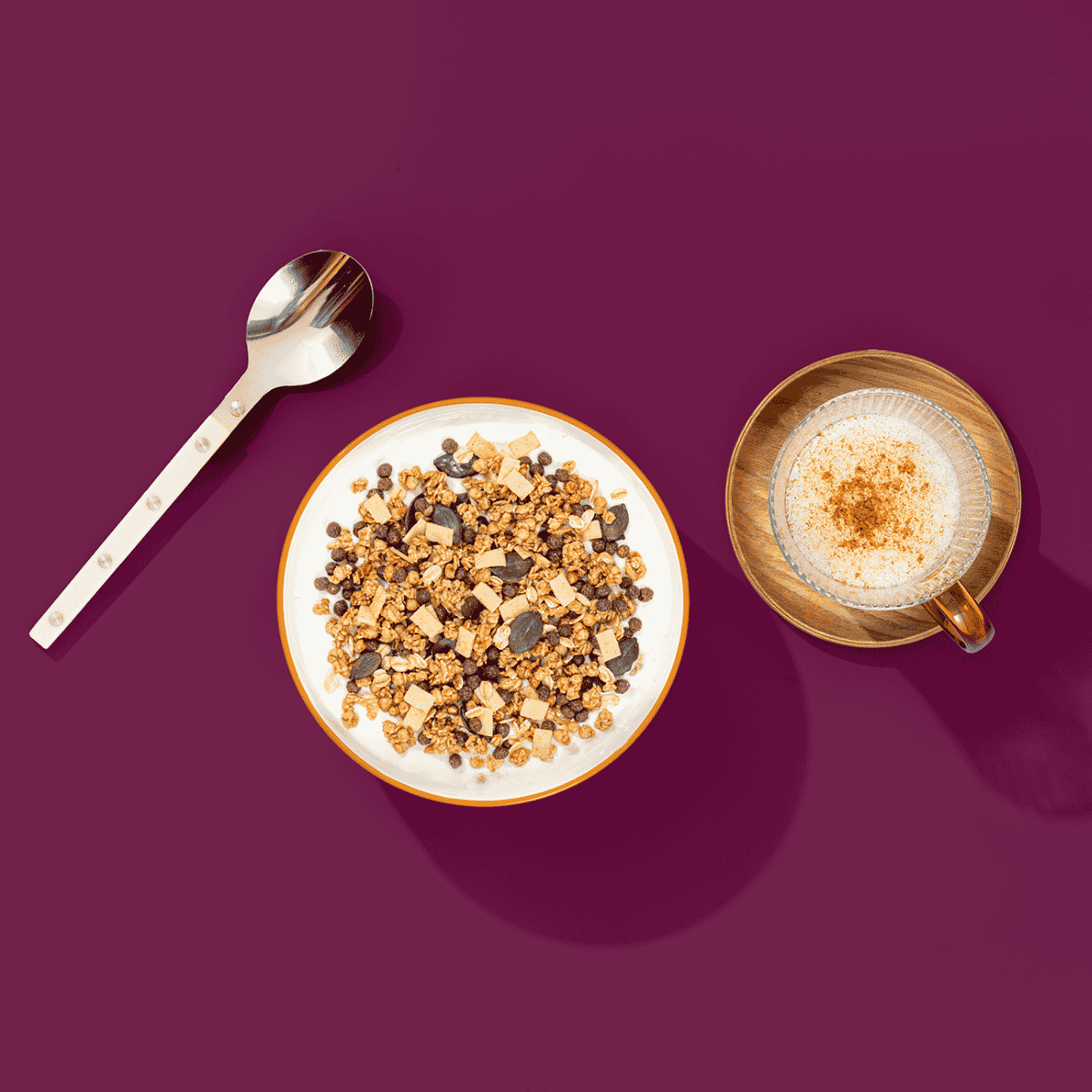Eine Schüssel mit Müsli. Nebendran ein silberner Löffel und ein Kaffee auf einem Holzuntersetzer.