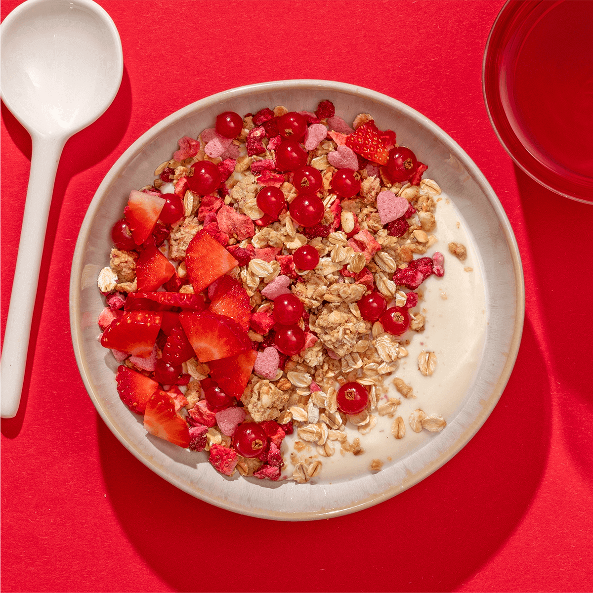 Nahaufnahme eines Müslis mit Beeren und Erdbeeren, links ein großer Löffel auf einem roten Hintergrund. 