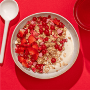 Nahaufnahme eines Müslis mit Beeren und Erdbeeren, links ein großer Löffel auf einem roten Hintergrund. 