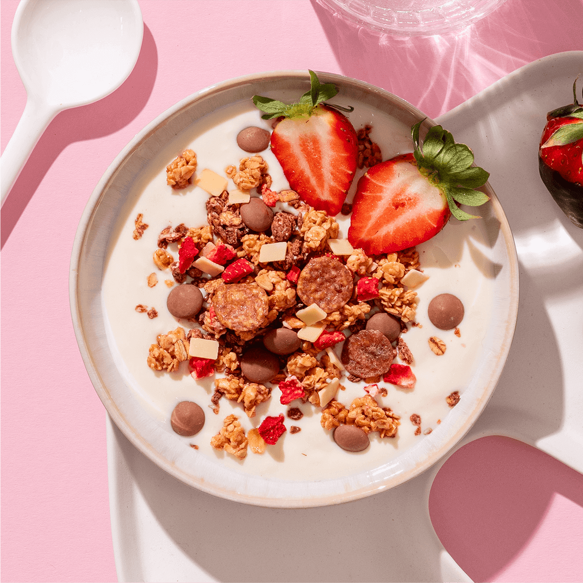 Eine Müsli-Bowl mit Joghurt, Schokostücken und frischen Erdbeerhälften auf organisch geformtem weißem Geschirr vor rosa Hintergrund.