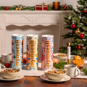 die drei Bio Weihnachtsmüslis  Choco Almond Cinnamon Crunch, Christmas Choco Aprocot Crunch und das Baked Apple Cookie Crunch stehen auf einer weihanachtlich gedeckten Tafel. IM Hintergrund ein Kamin und ein Weihnachtsbaum. 