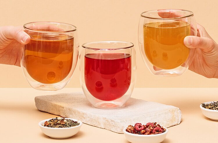 Drei transparente doppelwandige Teeglaeser mit verschiedenen Tees gefuellt neben Blatttees in kleinen Schaelchen